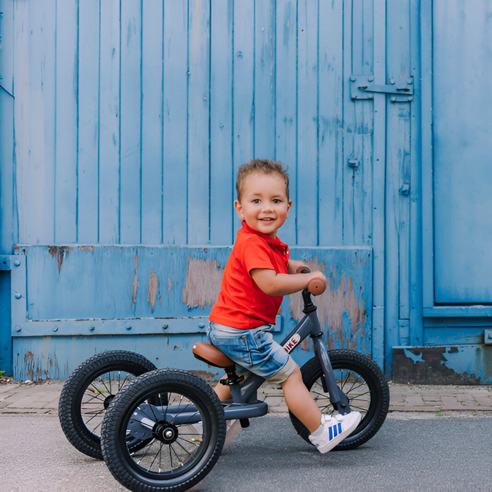 Little boy riding a Grey Trybike balance bike against a rustic blue wall.