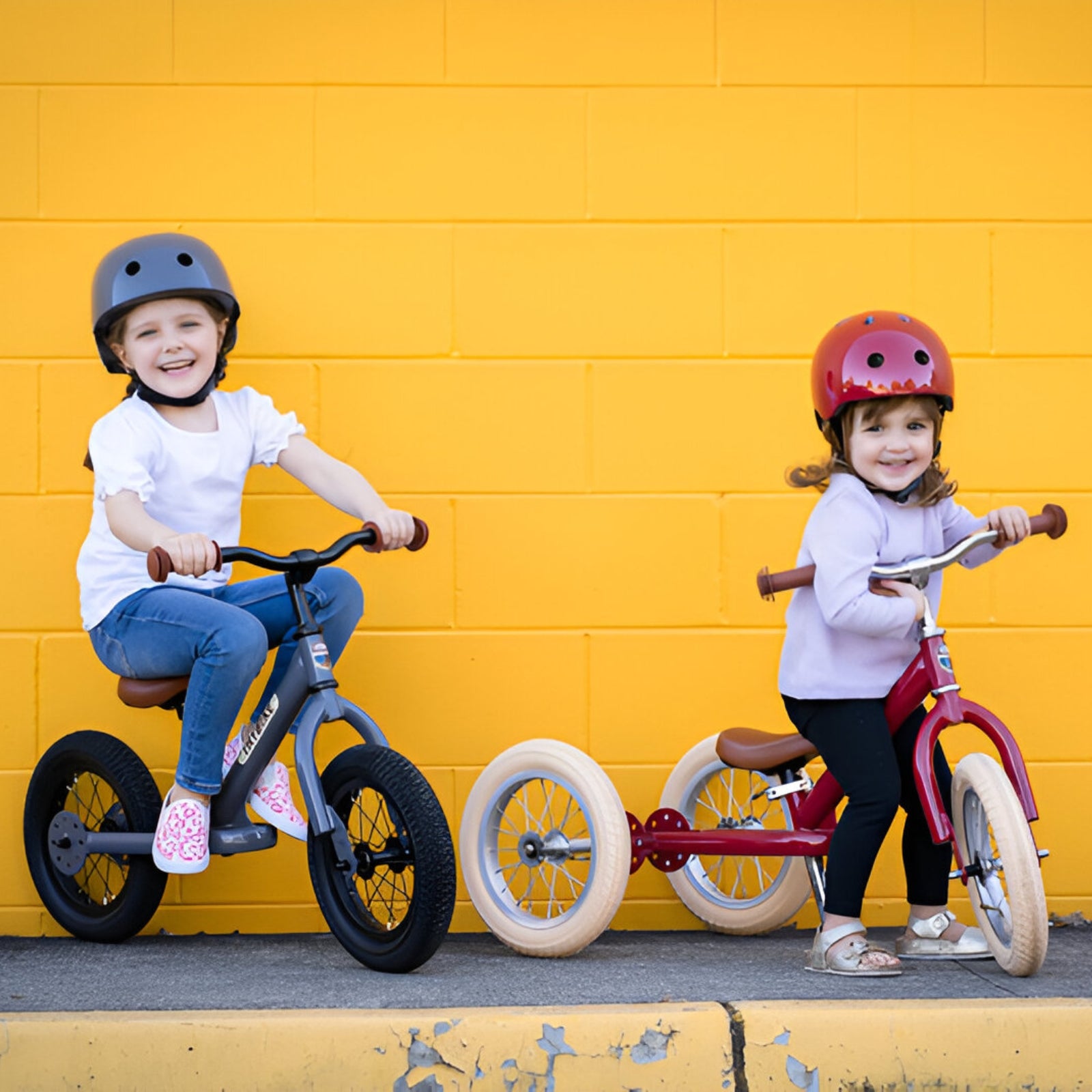 Two smiling kids sitting on their Grey Trybike and vintage red trybike in front of a bright yellow wall.