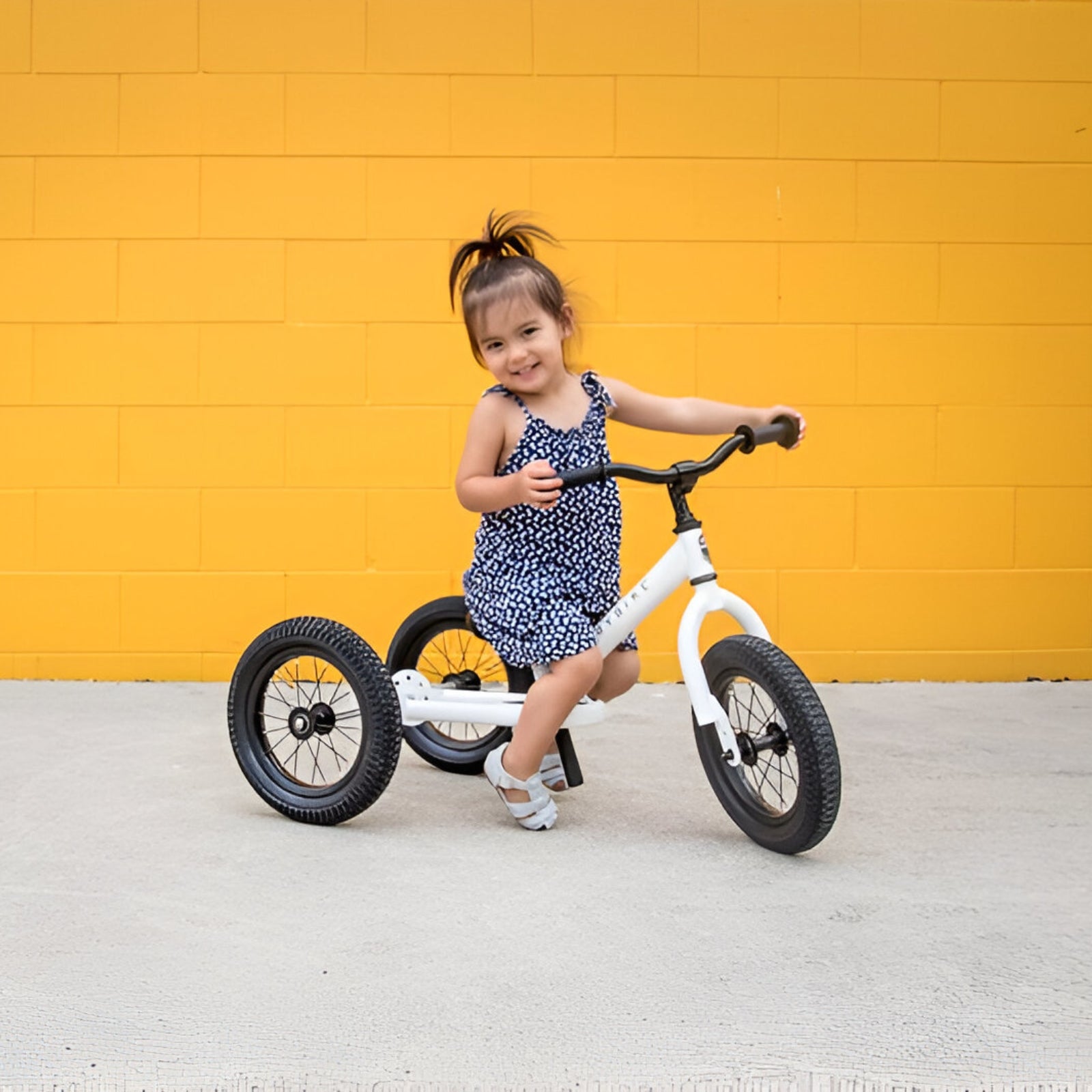 Happy toddler girl with a White Trybike on a vibrant yellow background.